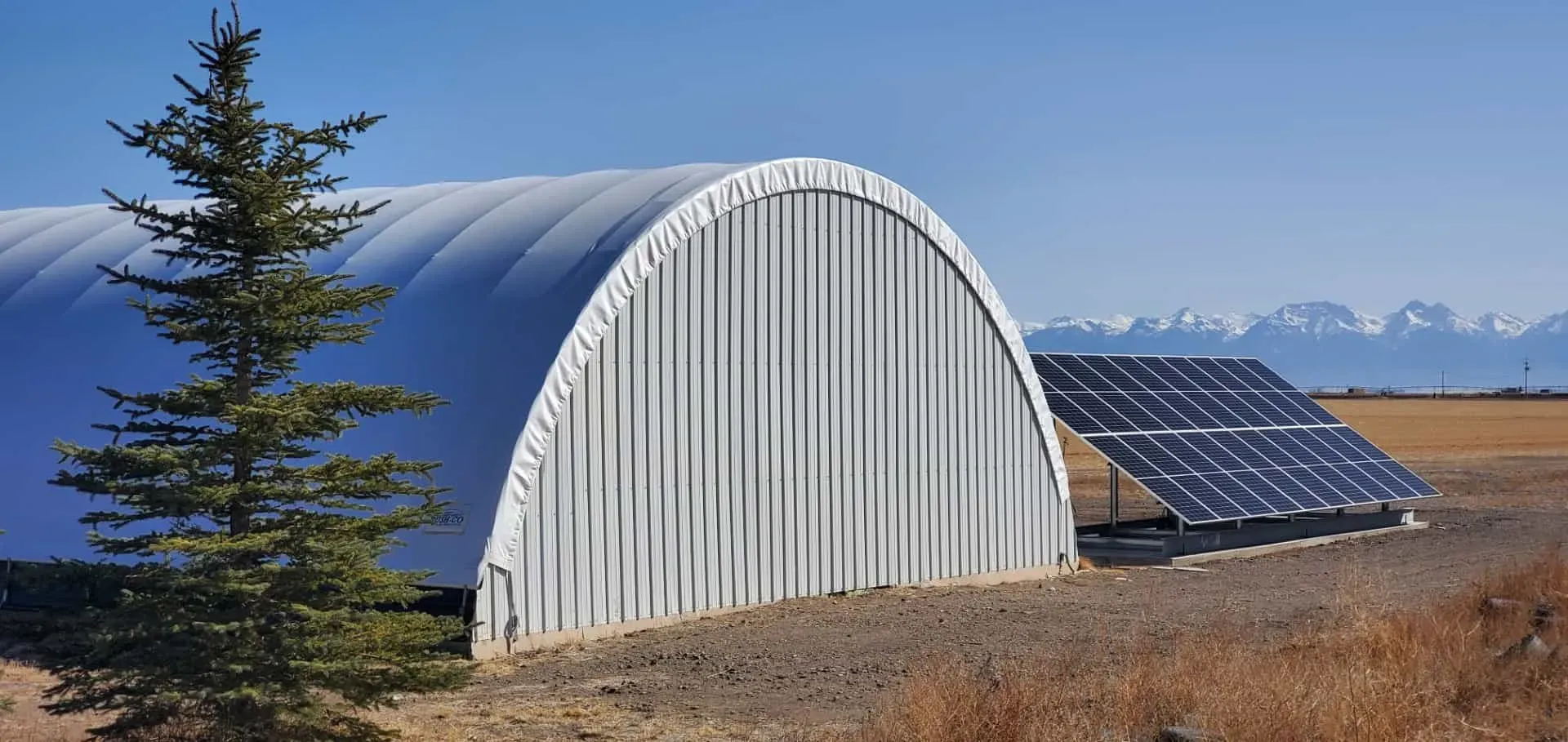Ground-mounted grid-tie solar array beside a farm building, with mountains in the distance